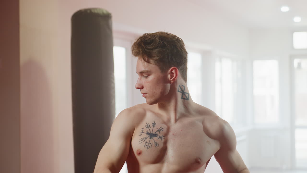 Man in black athletic shirt standing in bright gym interior with calm thoughtful posture, looking downward with focus, showing discipline, introspection, strength, and determination before workout