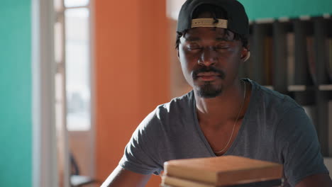 Profile view of man turning with glance, wearing cap backward in library, teal and orange walls soft light on face, faint stack of books in foreground thoughtful mood and slight tilt