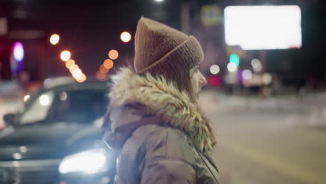 first-person view of a woman in a winter coat with a fur hood, crossing the road at night. blurry city lights, people, and cars are visible in the background