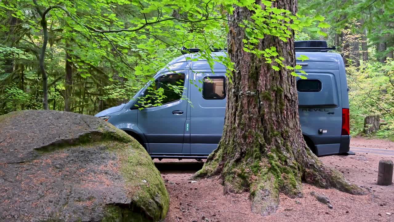 Cinematic drone shot of a camper van parked in a lush forest clearing surrounded by mossy trees and boulders