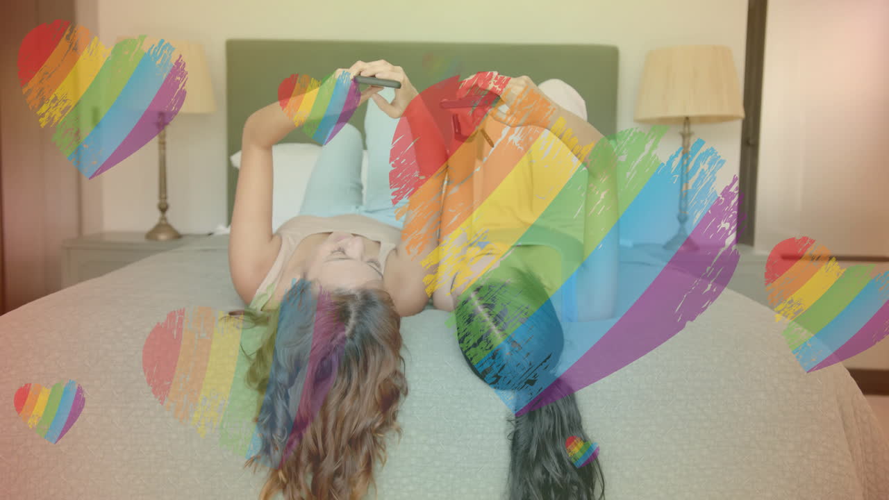 animación de corazones de arco iris sobre dos personas acostadas en la cama usando teléfonos inteligentes