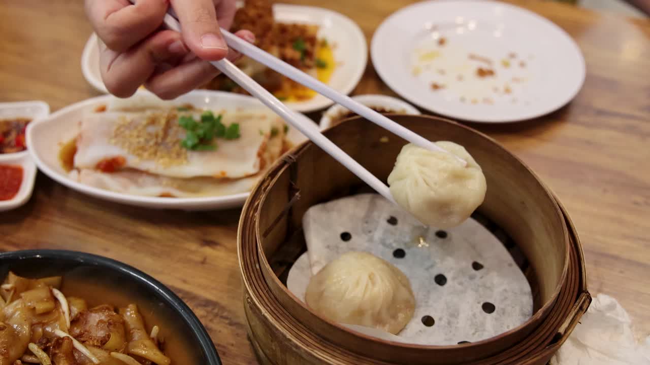 A hand uses chopsticks to lift a steamed xiao long bao dumpling from a bamboo basket in a brightly lit Singapore restaurant, surrounded by local dishes