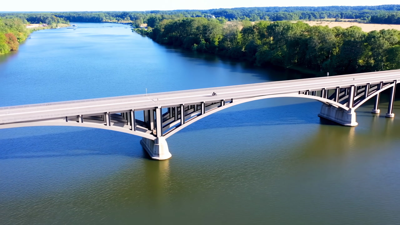 Aerial Bridge Over Calm River Showcasing Concrete Arch Spanning Wide Blue Water, Sunlit Roadway Serving