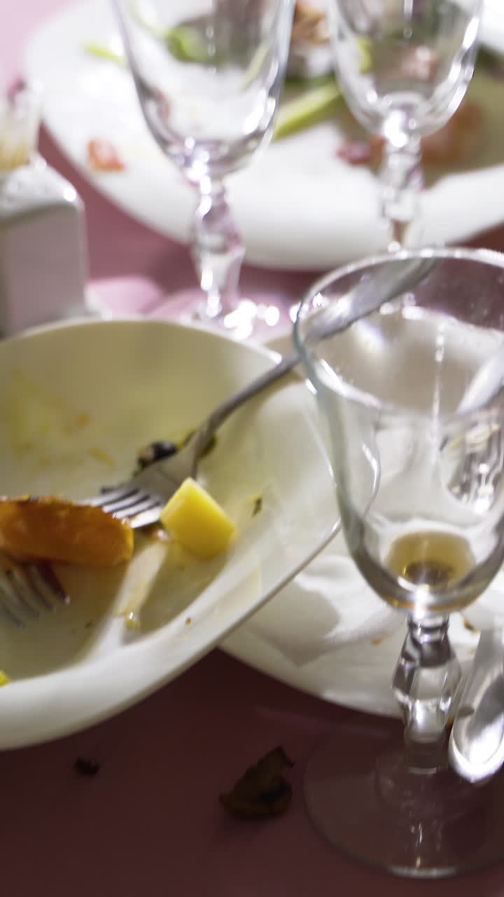 dirty plates and cutlery with wineglasses are located on a pink tablecloth. Camera motion forward Vertical video
