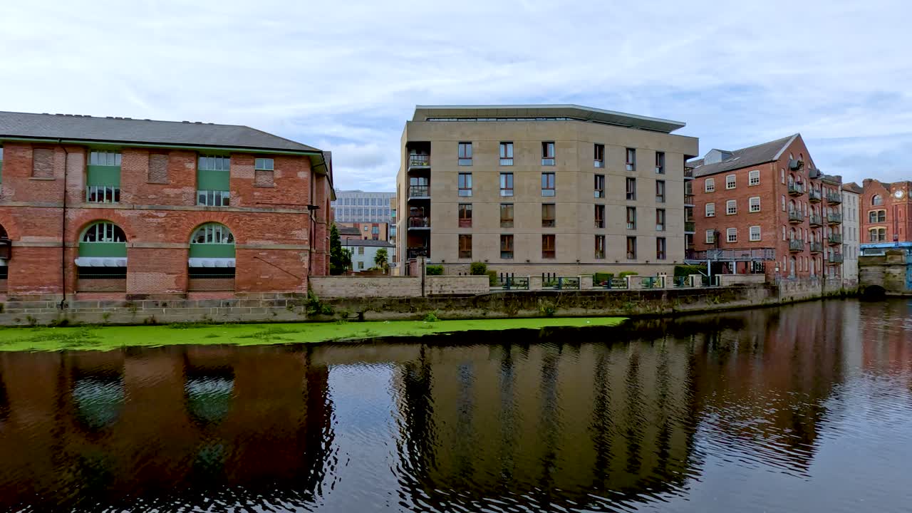 A steady daytime pan follows a calm river lined with historic brick buildings and modern apartments under a cloudy sky in Leeds, United Kingdom