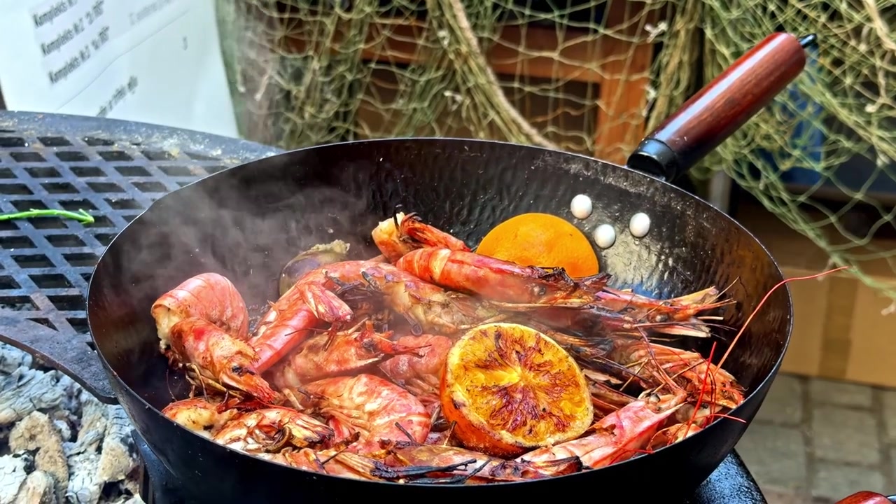 Large shrimp and citrus slices sizzling in a pan over an outdoor grill, with steam rising