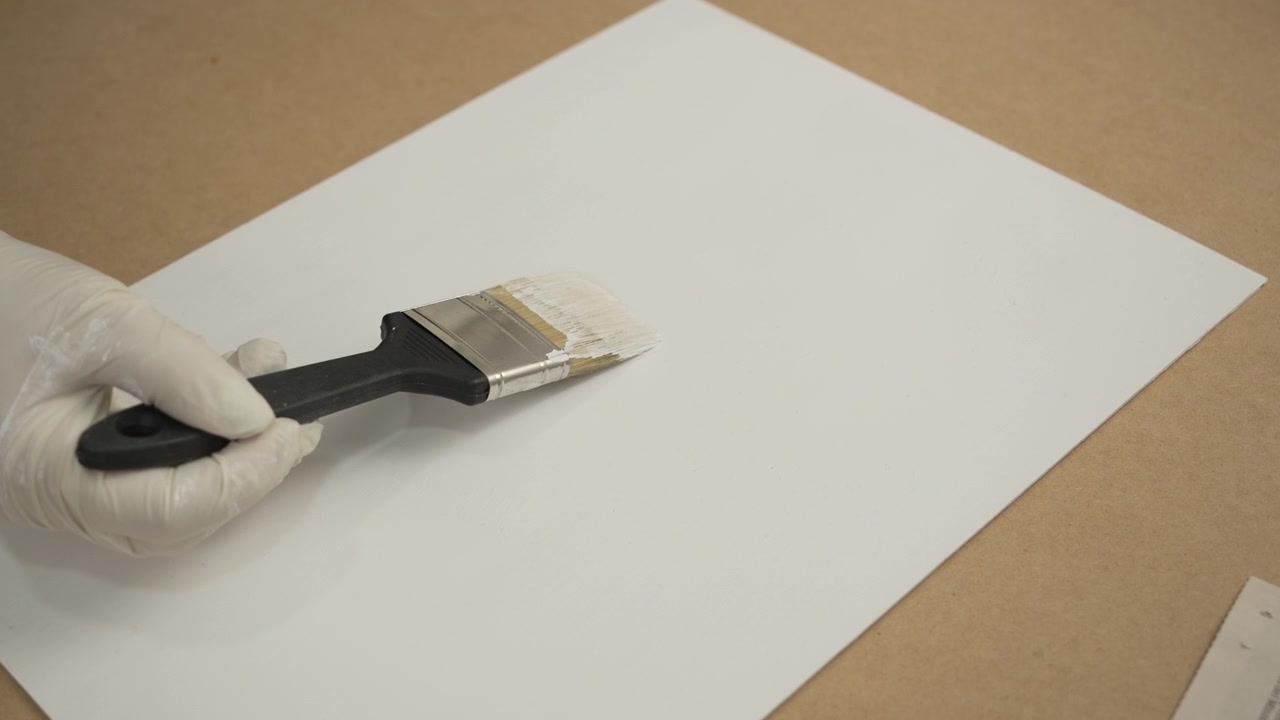 Mano guantata che utilizza un pennello per applicare vernice bianca o primer su un foglio di carta o un pannello