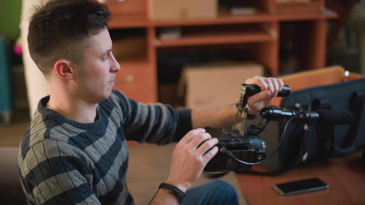Uomo che regola una videocamera su treppiede, preparando l'attrezzatura in uno spazio di lavoro creativo. Telefono e attrezzatura fotografica sul tavolo, mobili moderni con parete verde sullo sfondo, allestendo per riprese o creazione di contenuti.