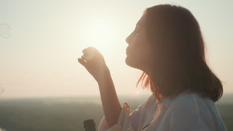 side view of woman holding bubble wand and blowing bubbles against glowing sunset sky with soft breeze in her hair, peaceful expression, dreamy golden light, distant forest landscape in background
