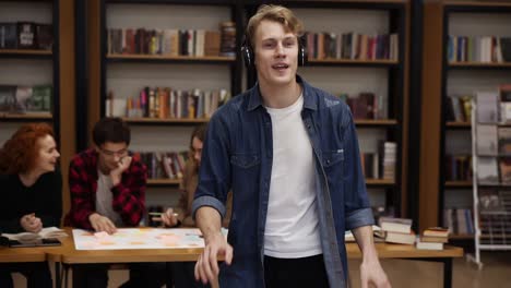 Handsome,-excited-european-man-in-denim-shirt-performing-expressive-dance-while-listening-favourite-music-in-headphones-in-academic-library-against-bookshelves-and-classmates-background.-Slow-motion