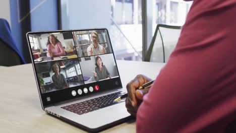 hombre afroamericano usando una computadora portátil para una llamada de video, con diversos colegas de negocios en la pantalla
