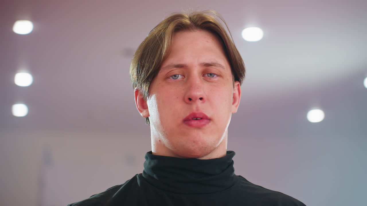 Fellow in black turtleneck stares with serious expression under bright lights, atmosphere filled with intensity and determination, focus, confidence, and mental discipline in indoor training space