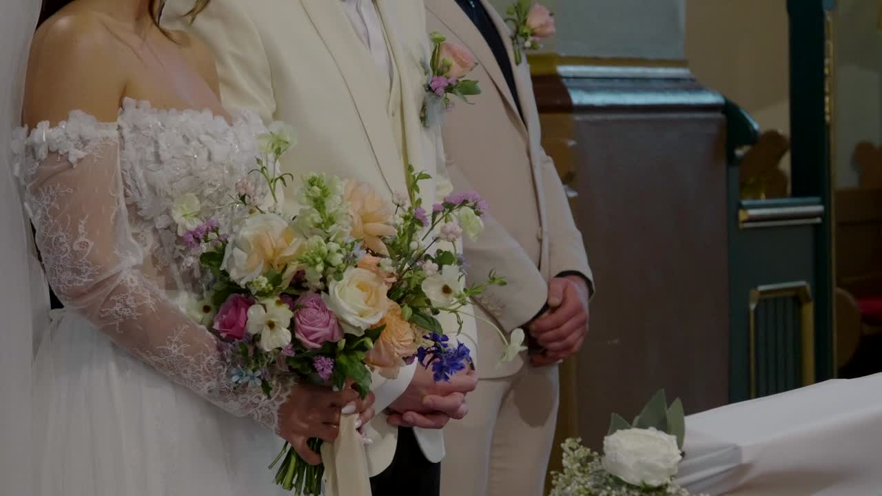la novia y el novio, vestidos con trajes de boda, se paran frente al altar en la iglesia. la novia sostiene un ramo de flores de colores