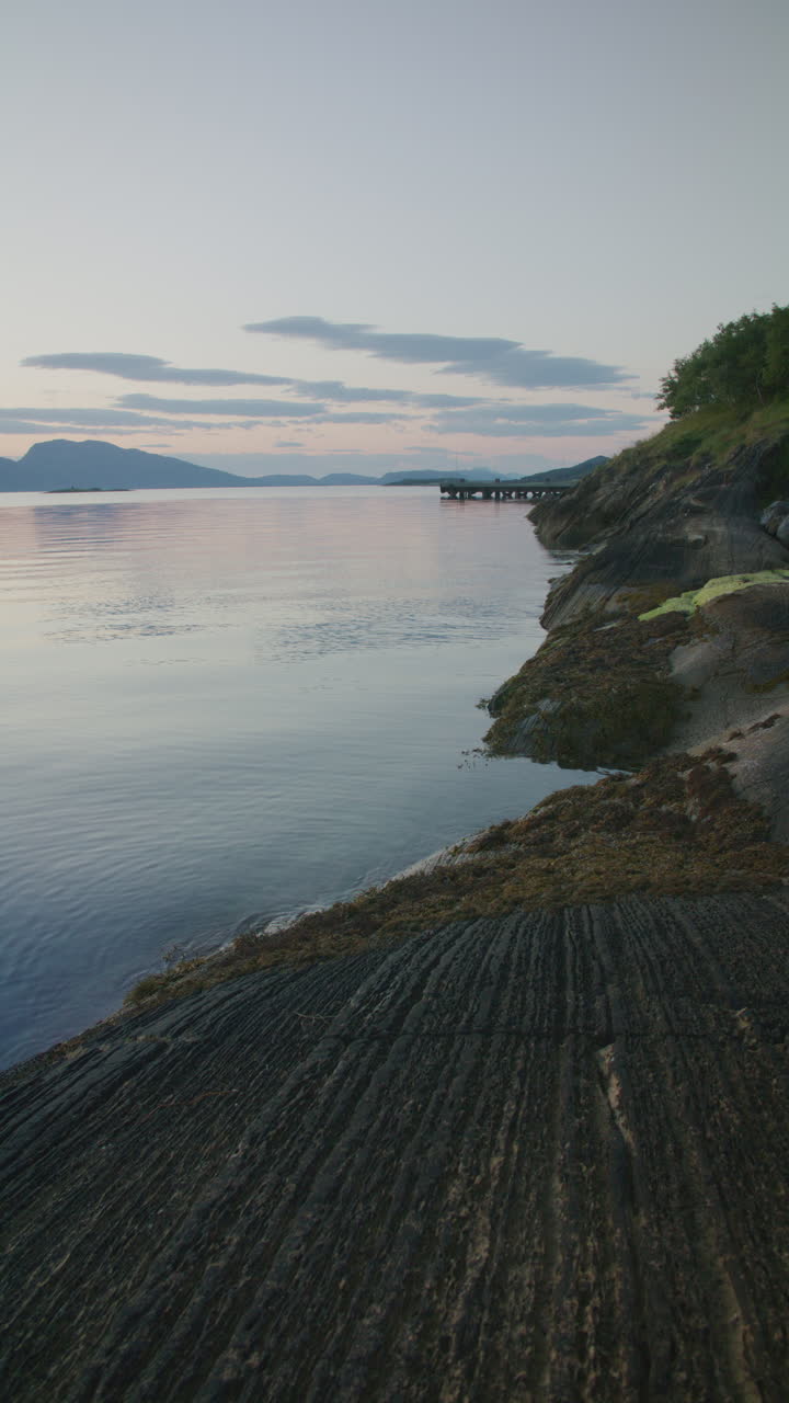 A vertical shot of a rugged shoreline with smooth water and a pier in the distance. Beautiful rock pattern filling the foreground. Captured in Northern Norway.
