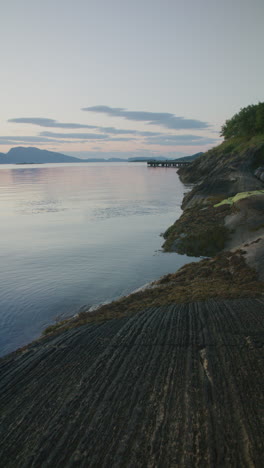 a vertical shot of a rugged shoreline with smooth water and a pier in the distance. beautiful rock pattern filling the foreground. captured in northern norway.