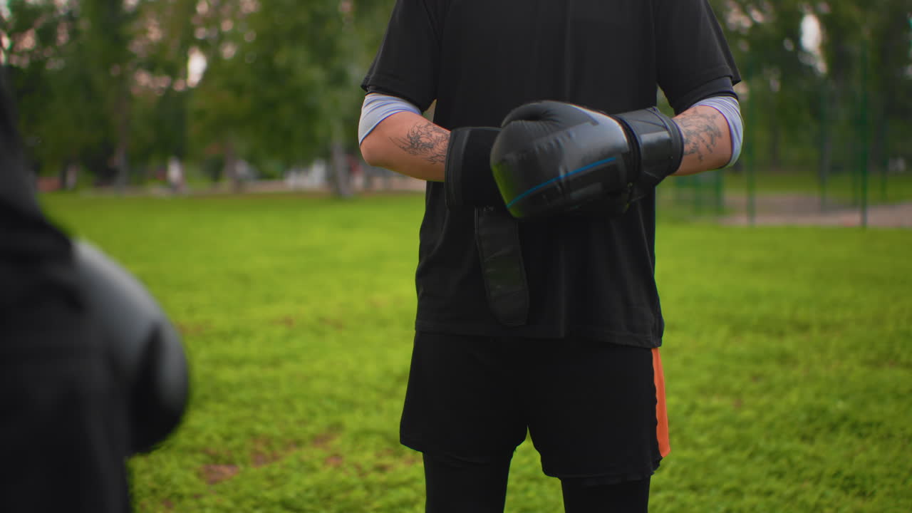 Closeup of white boxer wrapping gloves, intense handwrap ritual, visible forearm tattoos, black gloves and tightening straps, trainer mitt blurred foreground, dusk park background, tactile texture
