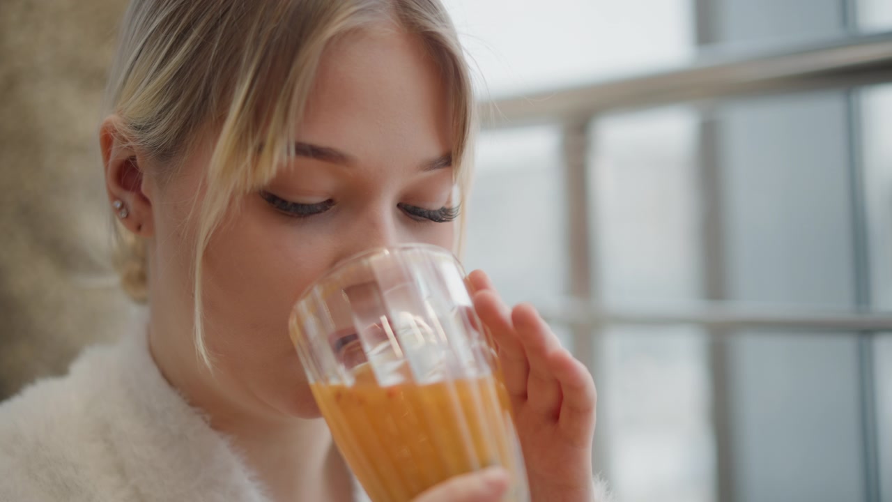 Mellomnærbilde av ung kvinne som fører et glass appelsinjuice til leppene og nipper med øynene delvis lukket, hodet vippet litt fremover og hånden hevet i en forsiktig drikkebevegelse i mykt innendørs lys