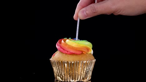 Hand places birthday candle in rainbow frosted cupcake with gold wrapper on black studio background