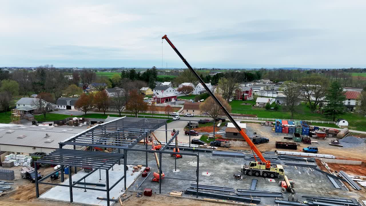 A construction site is busy with workers coordinating activities. A large crane is lifting materials to build a new structure. The setting is surrounded by houses and farmland on a cloudy day