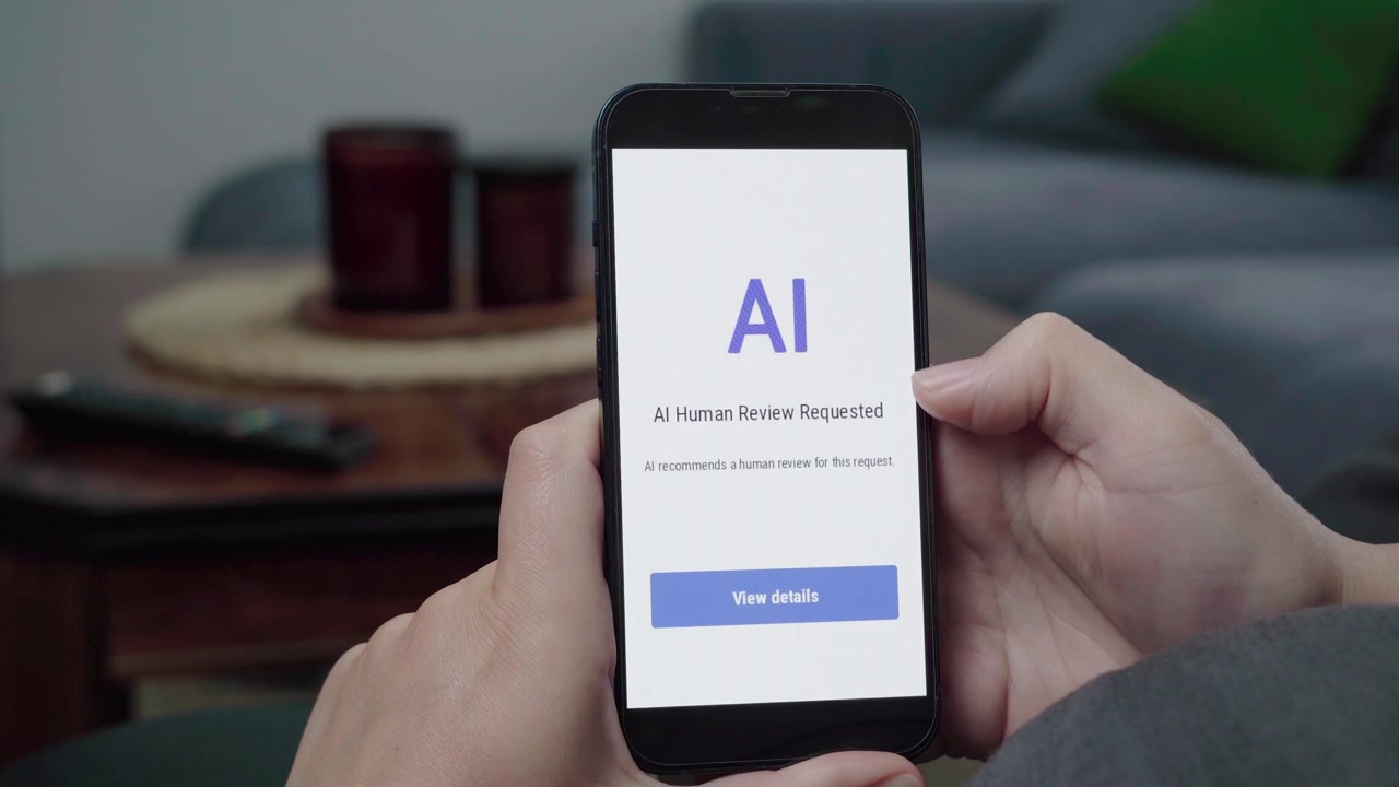 Mani usano un'app del telefono che mostra un avviso "Revisione umana AI richiesta" con un pulsante blu "Visualizza dettagli" in un soggiorno informale.