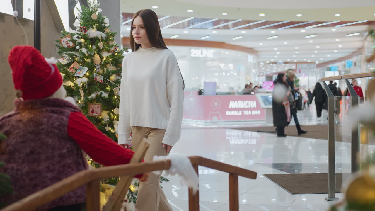 Lady walks up to decorative seat with festive plants in well-decorated mall with holiday decorations and people walking by, Christmas trees adorned with lights, holiday spirit in the air