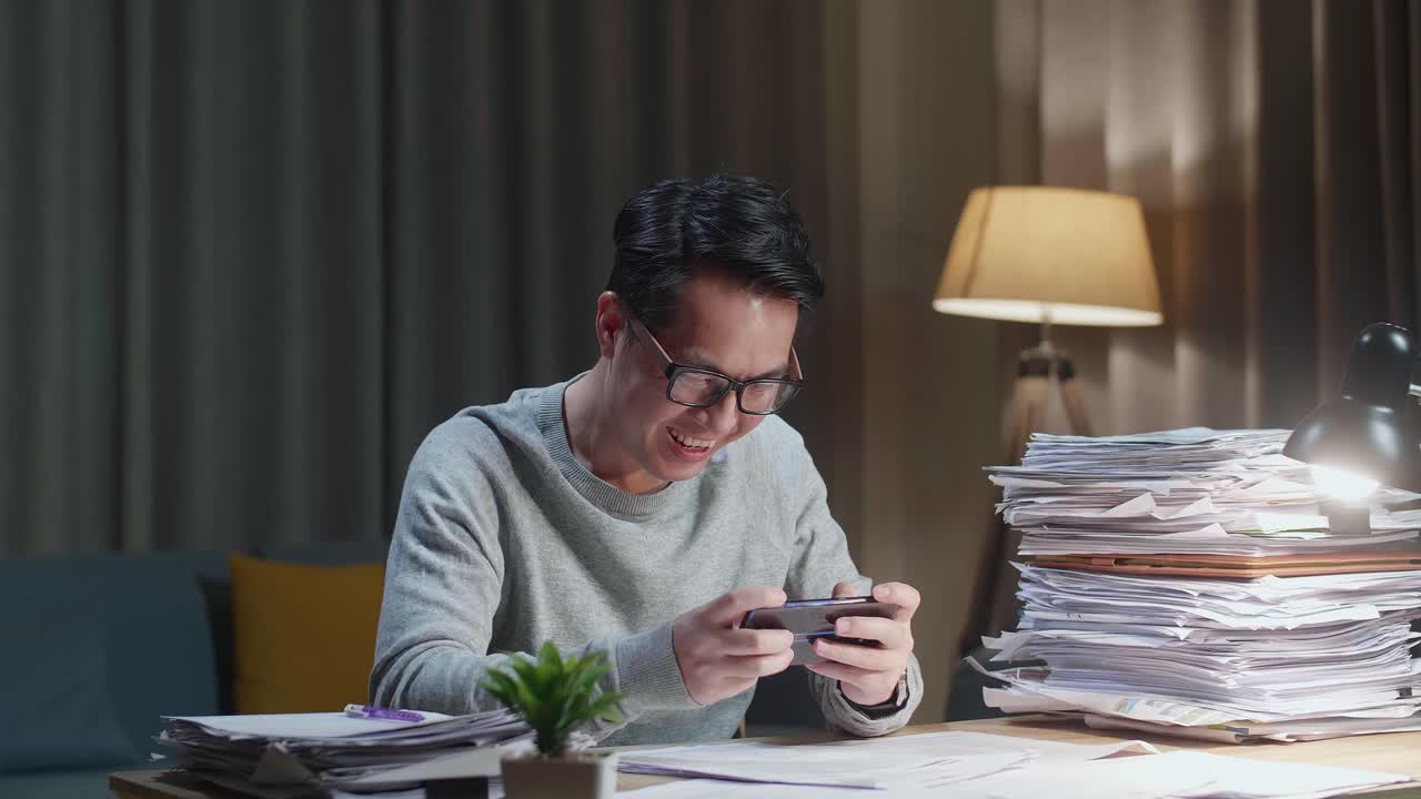 Happy Asian Man Celebrating Winning Game On Smartphone After Working With Documents At Home