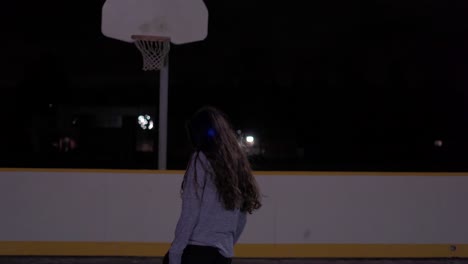 Una-Adolescente-Con-El-Pelo-Negro-Rebota-La-Pelota-Y-Luego-La-Dispara-En-La-Cancha-Al-Aire-Libre-Con-Un-Suéter-Gris-Por-La-Noche