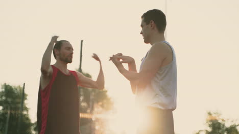 Jugadores-Masculinos-De-Baloncesto-Estirando-Los-Brazos-Y-Los-Hombros-En-Una-Cancha-De-Baloncesto-Al-Aire-Libre-Mientras-Hablan-Entre-Ellos