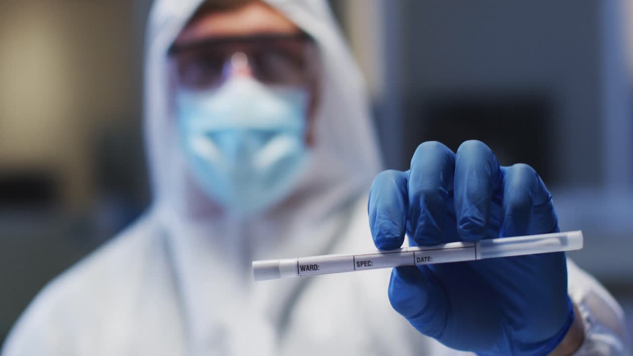Operatore sanitario caucasico di sesso maschile che indossa indumenti protettivi, mascherina e guanti, mentre tiene in mano un tampone di DNA in laboratorio.