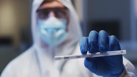 trabajador médico caucásico de sexo masculino con ropa de protección, máscara y guantes que sostiene un hisopo de adn en el laboratorio