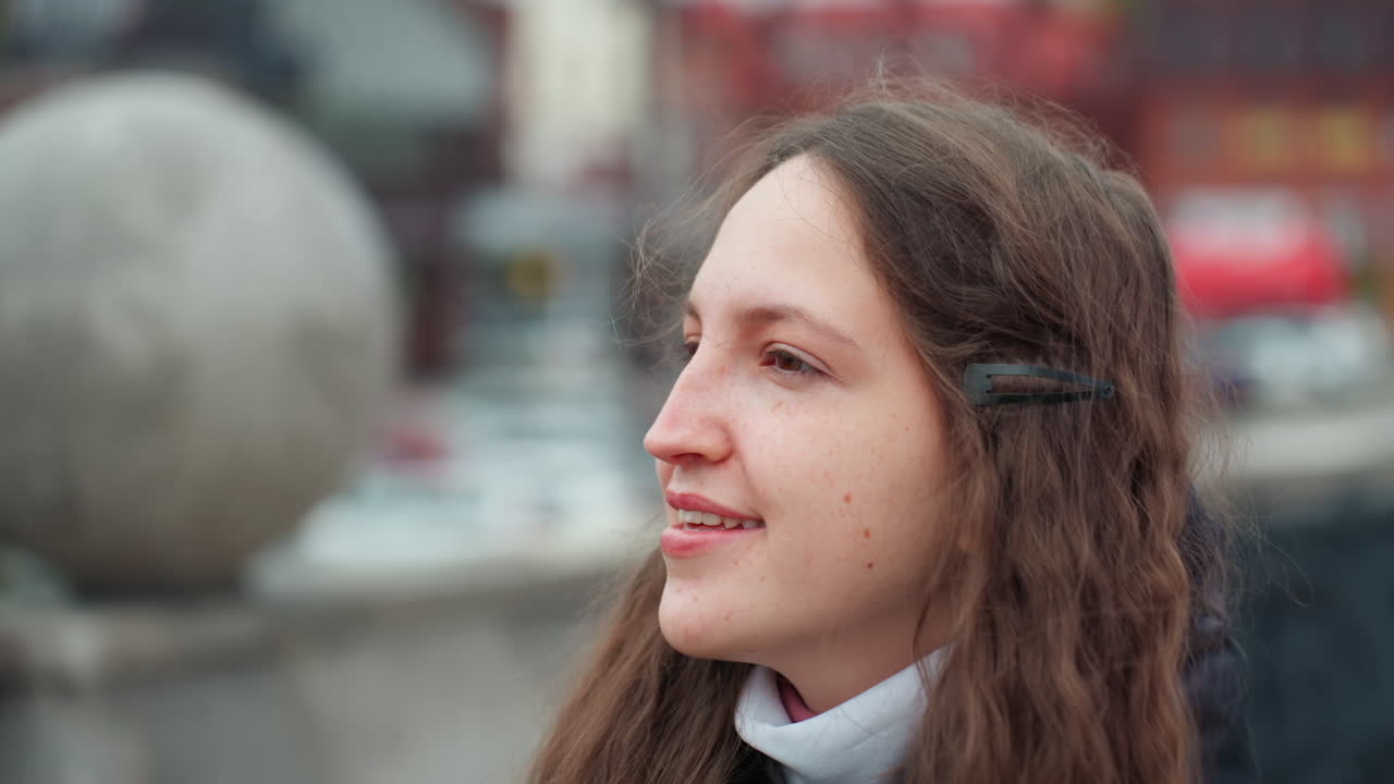 Donna caucasica che cammina per le vie della città, sorride e guarda verso l'alto, indossa un cappotto e una sciarpa, capelli lunghi sciolti, porta uno zaino, passa davanti a edifici colorati e a un ponte, cielo nuvoloso, passo rilassato