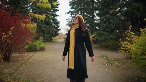 creative soul with wavy red hair adjusts her yellow beret while strolling through a lush garden, draped in a long yellow scarf, showcasing vibrant autumn foliage in the background