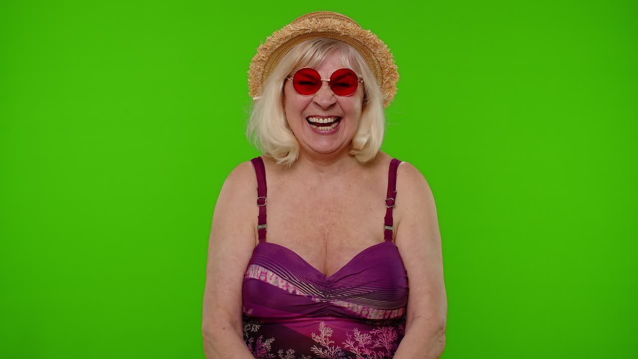 Anziana donna viaggiatrice in vacanza con occhiali da sole rossi, che guarda la telecamera, sorride e inizia a ridere