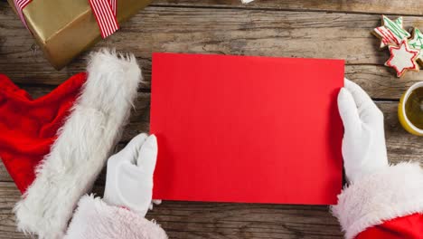 Animation-of-hands-of-santa-claus-holding-red-christmas-card-with-copy-space-over-wooden-background