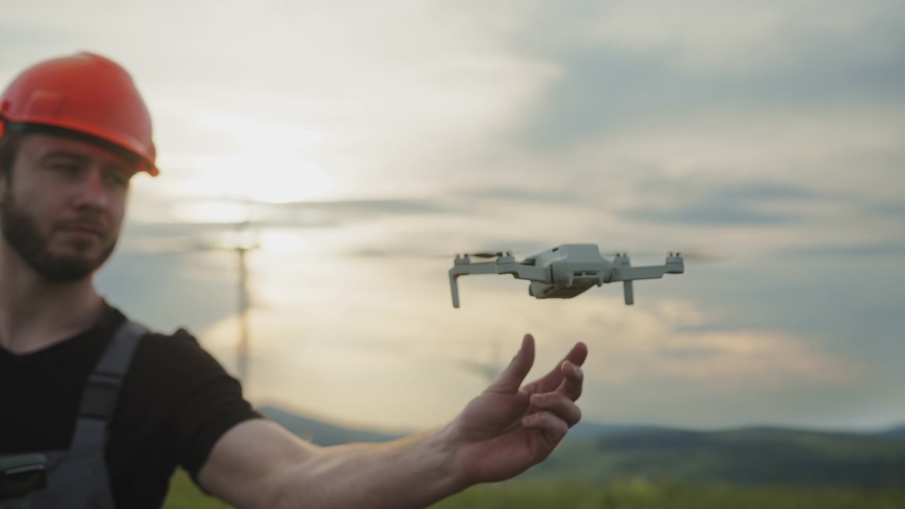 Piccolo drone bianco che sorvola colline verdi con pale eoliche in lontananza, prospettiva aerea