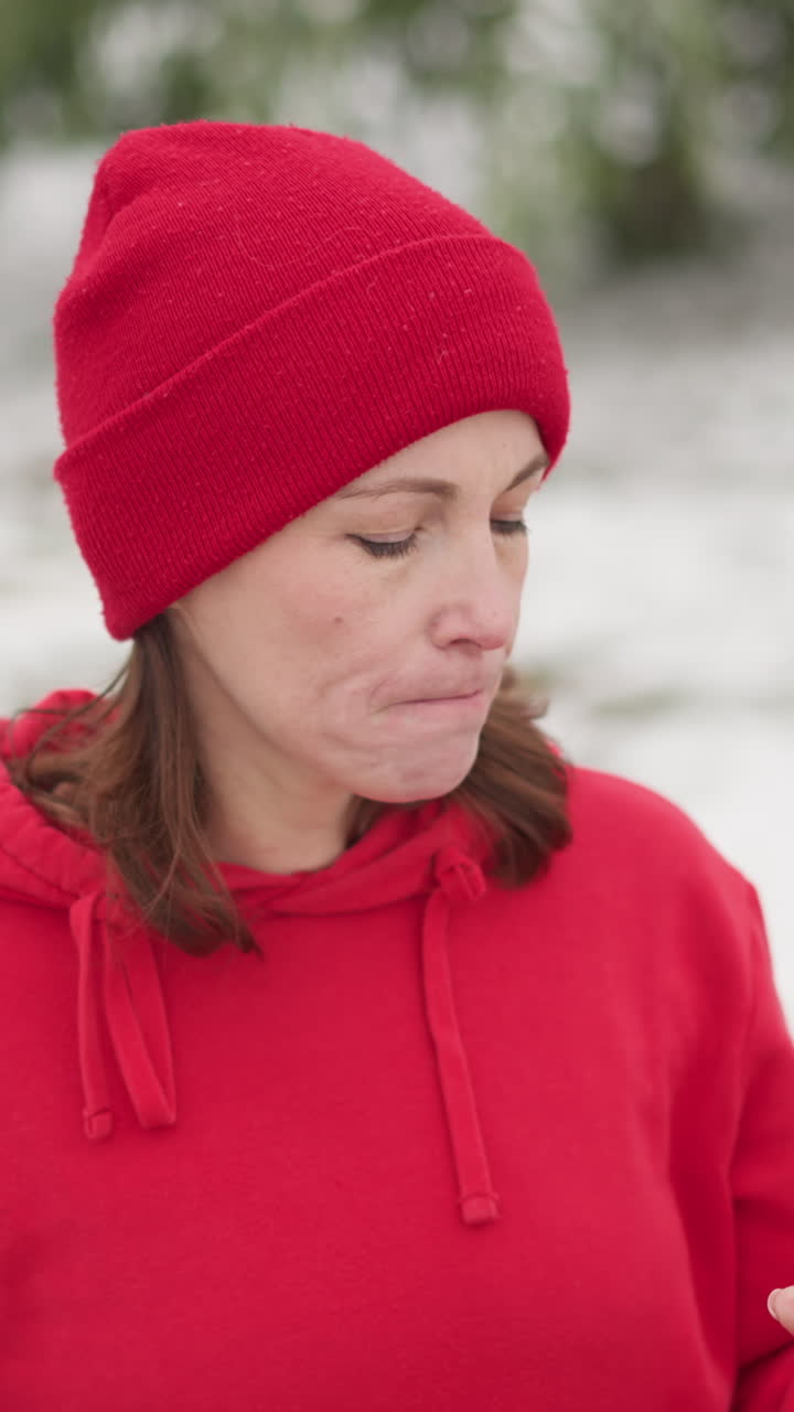 lady in red beanie and hoodie sipping water from pink bottle outdoors, blurred background showing bag on frosted bench, greenery, and iron rail in serene winter setting with light snow