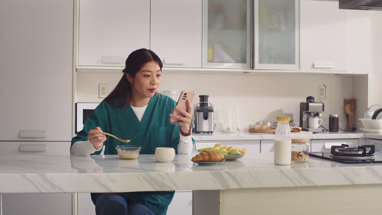 Asian Nurse Eating Yogurt and Using Phone at Kitchen