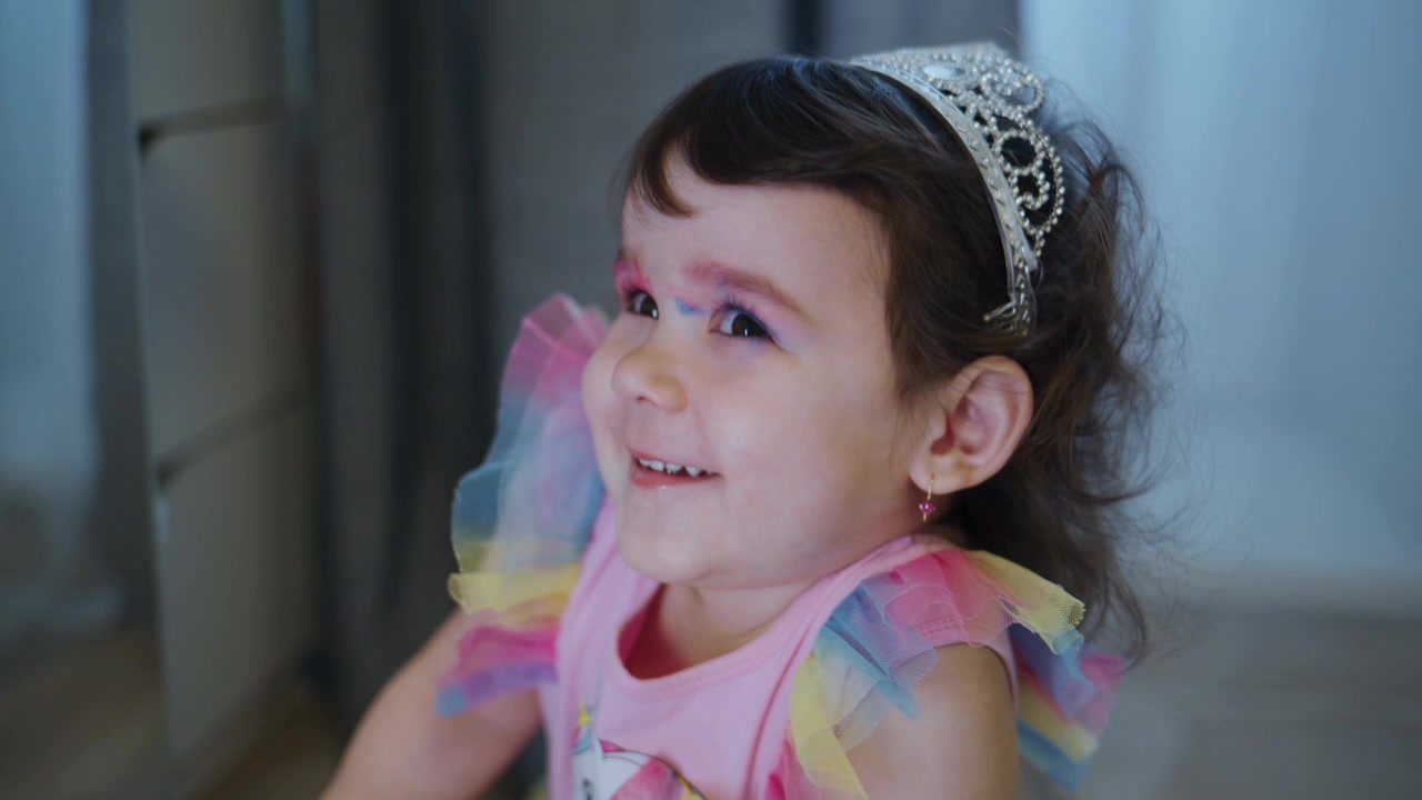 Young child wears tiara, colorful dress, and paint on face. Smiles broadly, eyes closed, clearly joyful. Hair messy, outfit festive, expression pure happiness. Perfect for birthday parties
