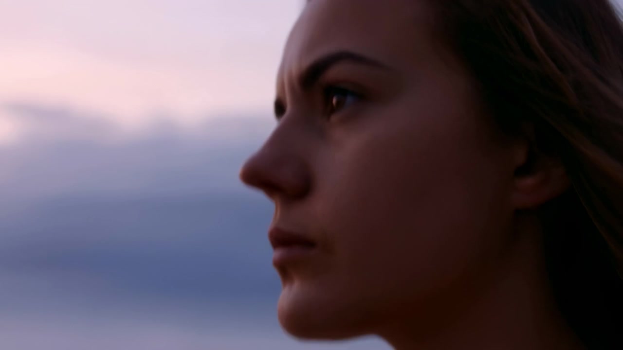 Young woman side profile beside calm ocean at dusk, with soft pink and blue sky, blurred horizon, gentle low light, smooth skin detail, and quiet reflective mood in tight close composition