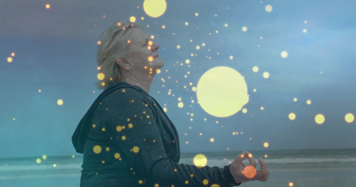 Senior woman meditating on shore for wellness, breath causing golden particles rising around her