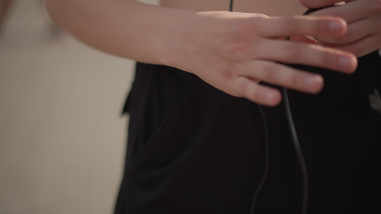 Penari muda melepaskan earbud yang kusut di jalan, close-up tangan yang berusaha memisahkan kabel yang kusut sebelum latihan di luar ruangan. Trotoar perkotaan yang diterangi cahaya matahari dengan crop top dan celana pendek kasual, fokus sebelum pertunjukan