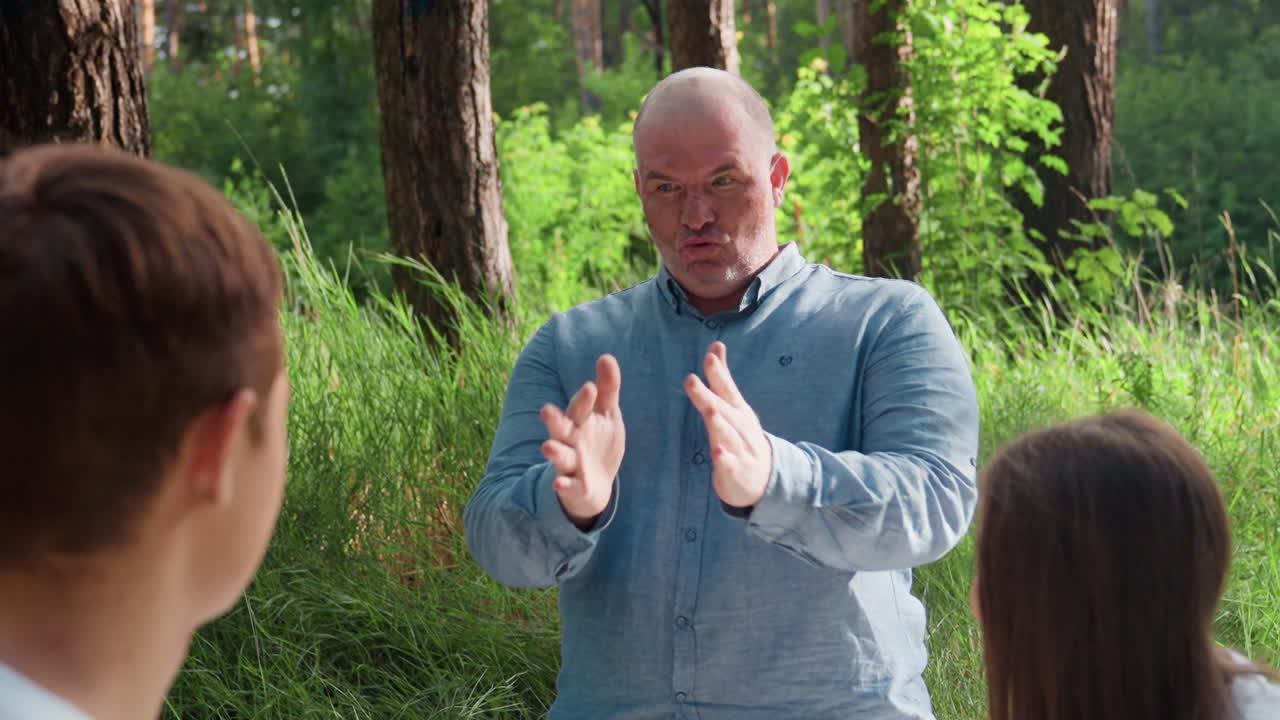 Head of family animatedly talking to kids during picnic surrounded by tall trees and sunlight, expressing emotions through gestures while children listen attentively, creating warm family connection