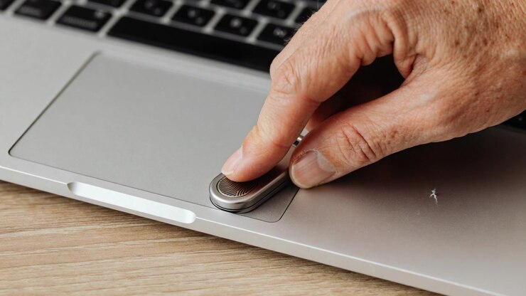 Una mano entra nell'inquadratura, posizionando un portachiavi decorato su un tappetino argentato per laptop su una scrivania di legno, sbloccando il laptop.