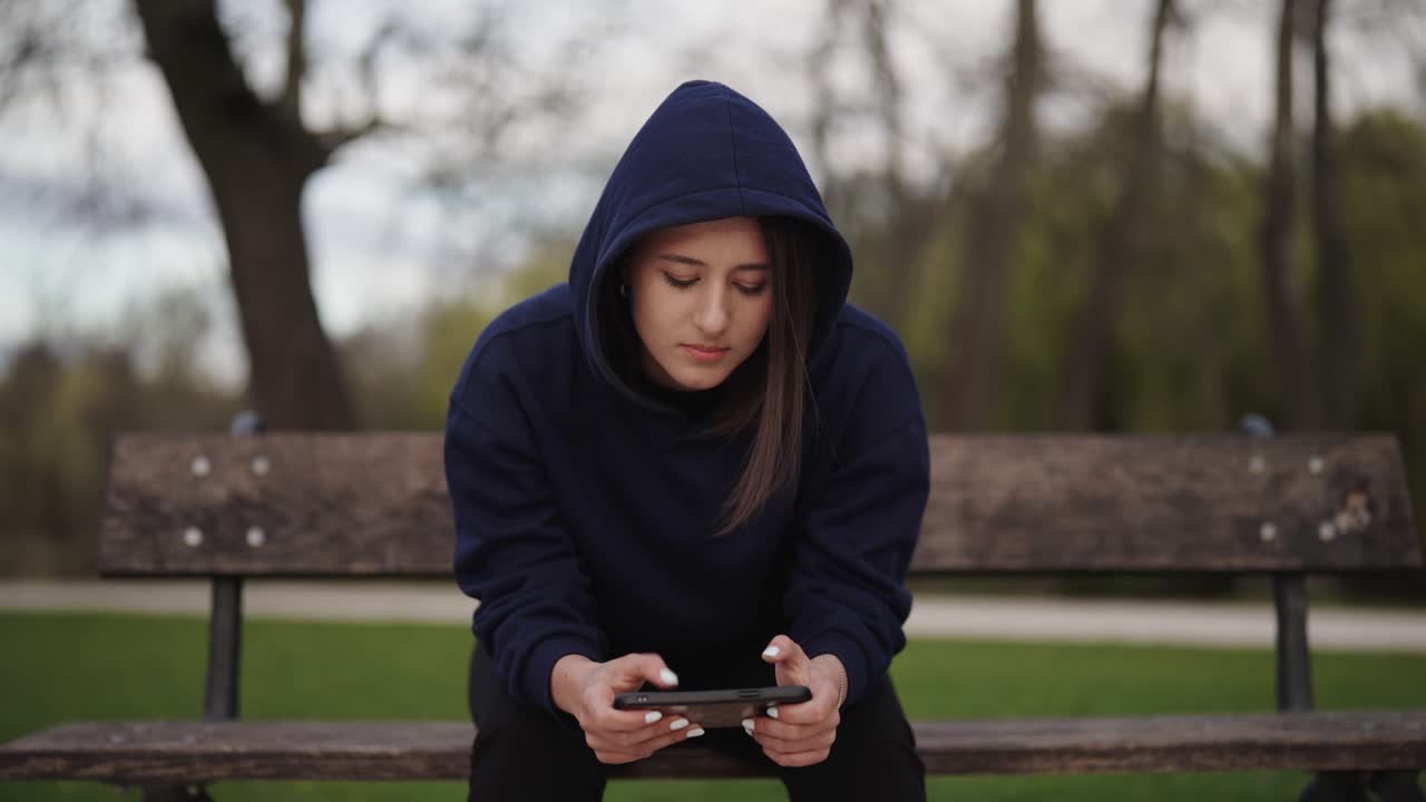 Donna solitaria con felpa con cappuccio seduta su una panchina di legno del parco e che usa lo smartphone, Praga