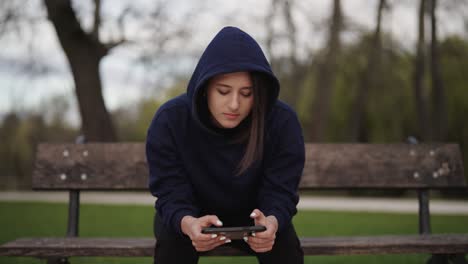 lonely woman in hoodie sit on wooden park bench and use smartphone, prague