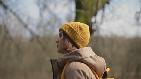 A-Young-Girl-In-A-Yellow-Wool-Cap-And-A-Yellow-Backpack-Walks-Through-The-Forest-Towards-The-Lake