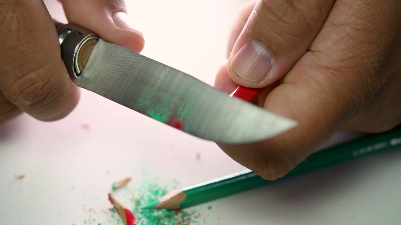 Footage of hands slowly sharpening a pencil and some coloured pencils with a sharp knife