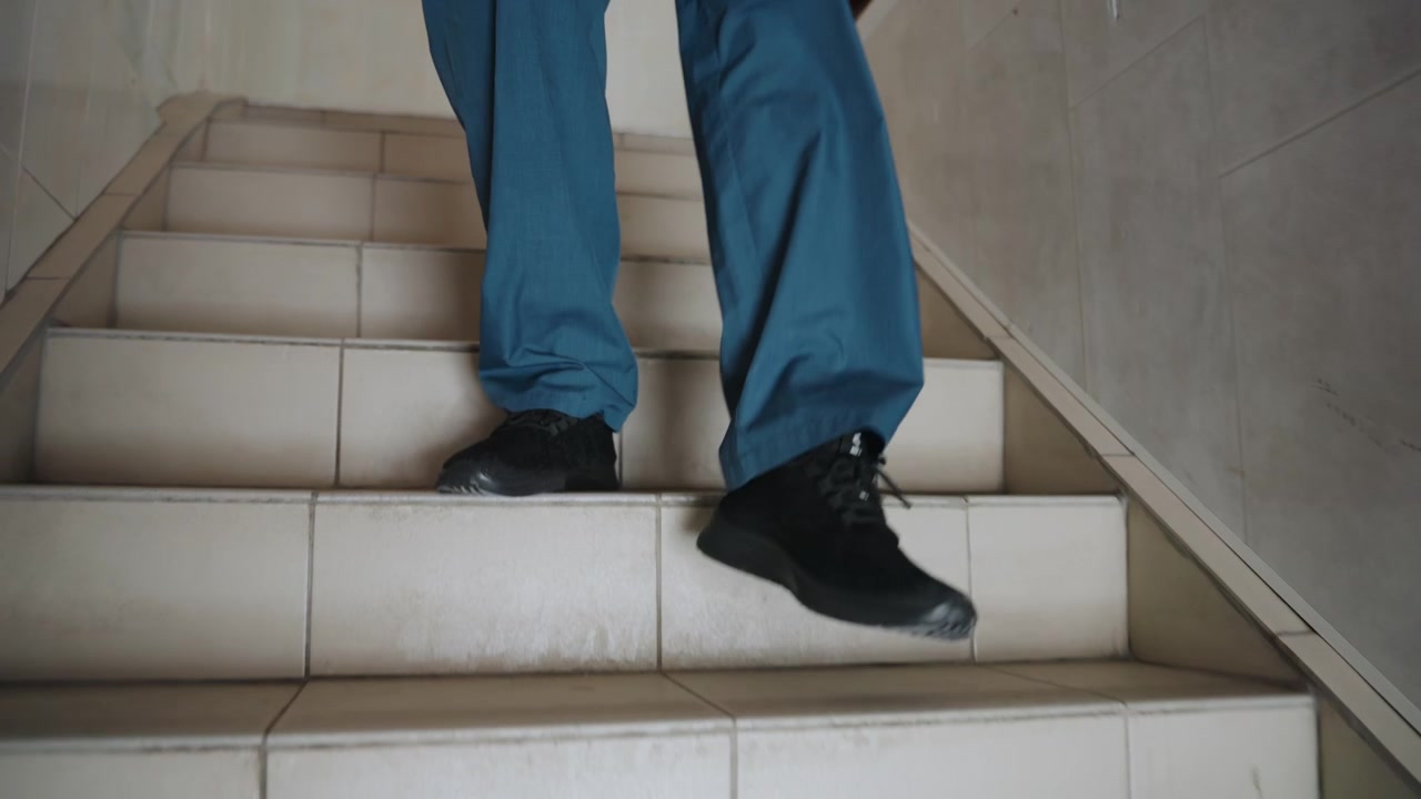 Scrub clad medic descending tiled stairs during urgent home call, steady footsteps on worn tiles, black shoes, blue scrubs, approaching apartment doorway, close up on lower body conveys tension