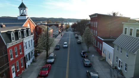 Drone-view-panning-upwards-of-a-main-street-in-small-town-America-at-sunset-with-reflections-off-of-the-homes-and-buildings-with-a-river-and-hilly-terrain-in-the-background