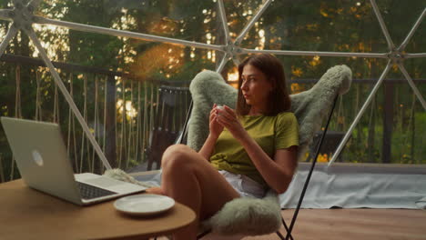 depressed woman drinks hot drink in silence working online on laptop with window view of forest at sunset. relaxation in solitude and peace with nature slow motion